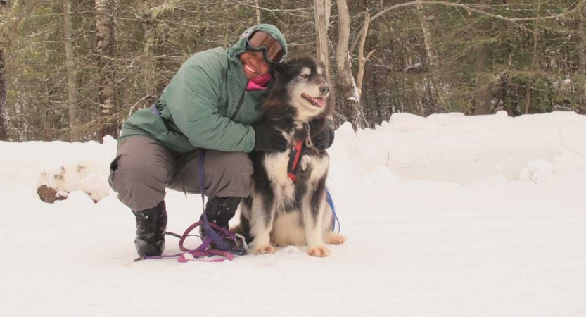 a person wearing snow gear smiles and bends down to hug a sled dog